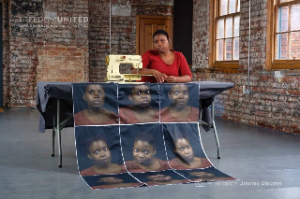 Woman sitting on a table with multiple pictures of her face printed on a poster that is in front of the table.