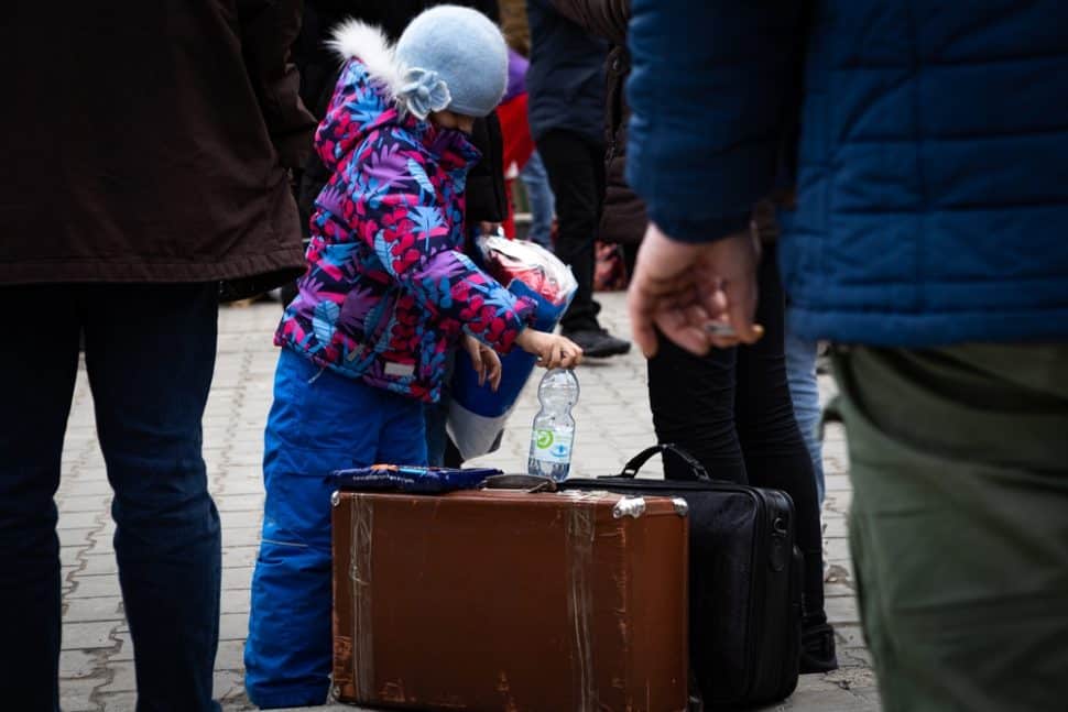 Girl dressed in winter coat and woolly hat with face hidden holding a water bottle looks down at suitcase at her feet whilst the legs of adults are around her.