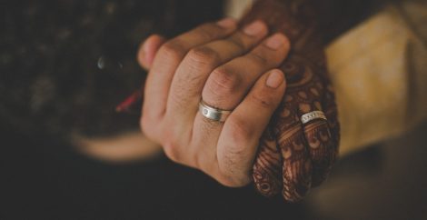 Dos manos abrazadas, ambas con anillos de boda. Uno de ellos tiene tatuajes de henna por todas partes.