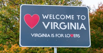 Blaues Schild mit der Aufschrift „Willkommen in Virginia: Virginia ist für Liebhaber“ hinter einem Hintergrund aus grünen Bäumen.