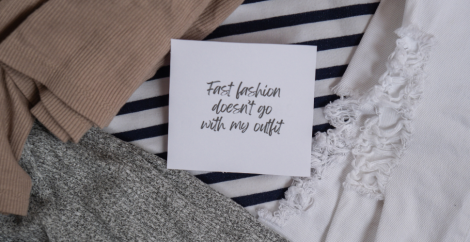 Zitat „Fast Fashion passt nicht zu meinem Outfit“ auf einem Zettel über verschiedenen Kleidungsstücken