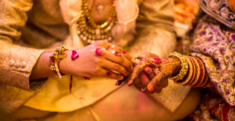 An Indian couple in traditional wedding attire, the man putting a ring on the woman's finger.