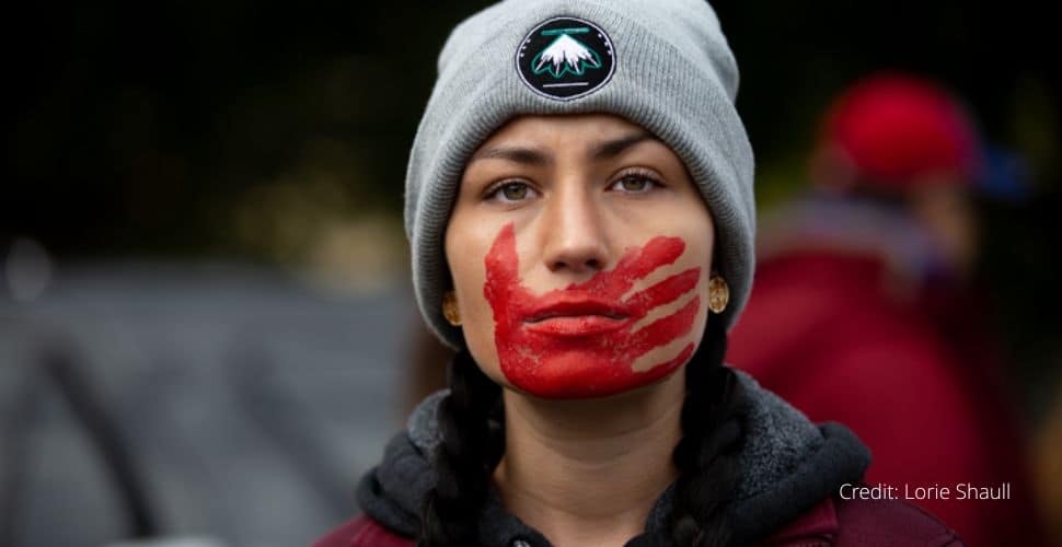 A headshot of a woman wearing a woollen hat with red paint across her mouth and lower face suggesting an imprinted hand.