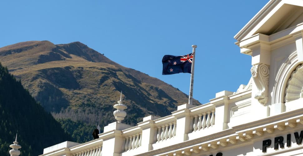 New Zealand flag blowing in the wind atop a pole on the roof of a white building against a backdrop of mountains