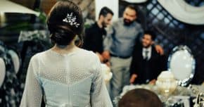 Eine Frau im Brautkleid steht mit dem Rücken zur Kamera, drei Männer unterhalten sich vor ihr.
